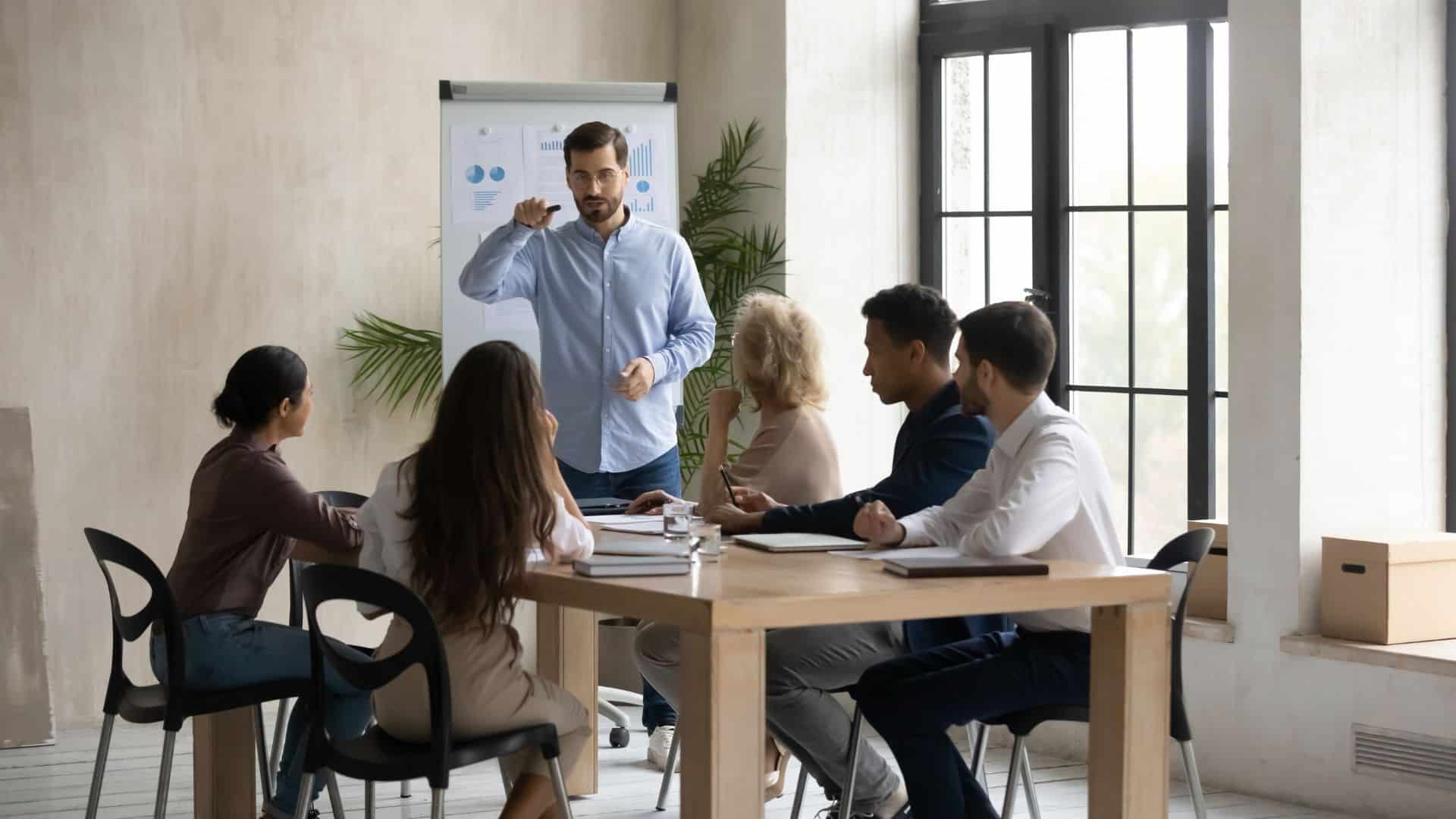 business coach standing in front of 5 team member seating in a table