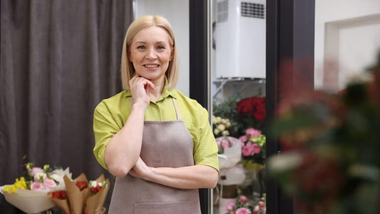Smiling female small business owner in a flower shop wearing an apron, representing confidence, work–life balance, and positive mental health among entrepreneurs.
